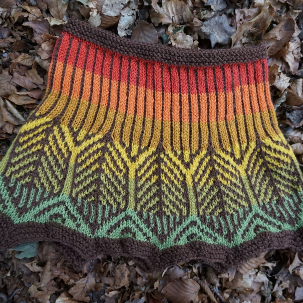 a cowl lying on leaves. It is created in mosaic knitting in fall colours.