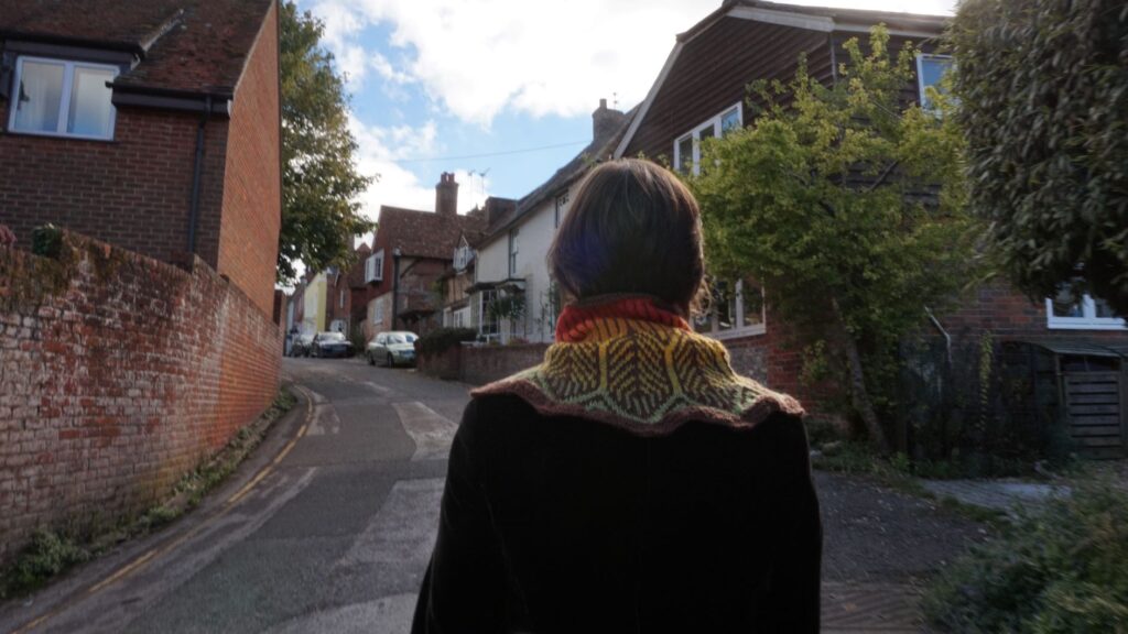 back view of a woman walking down a street with old houses on each side. she is wearing a cowl and brown jacket.