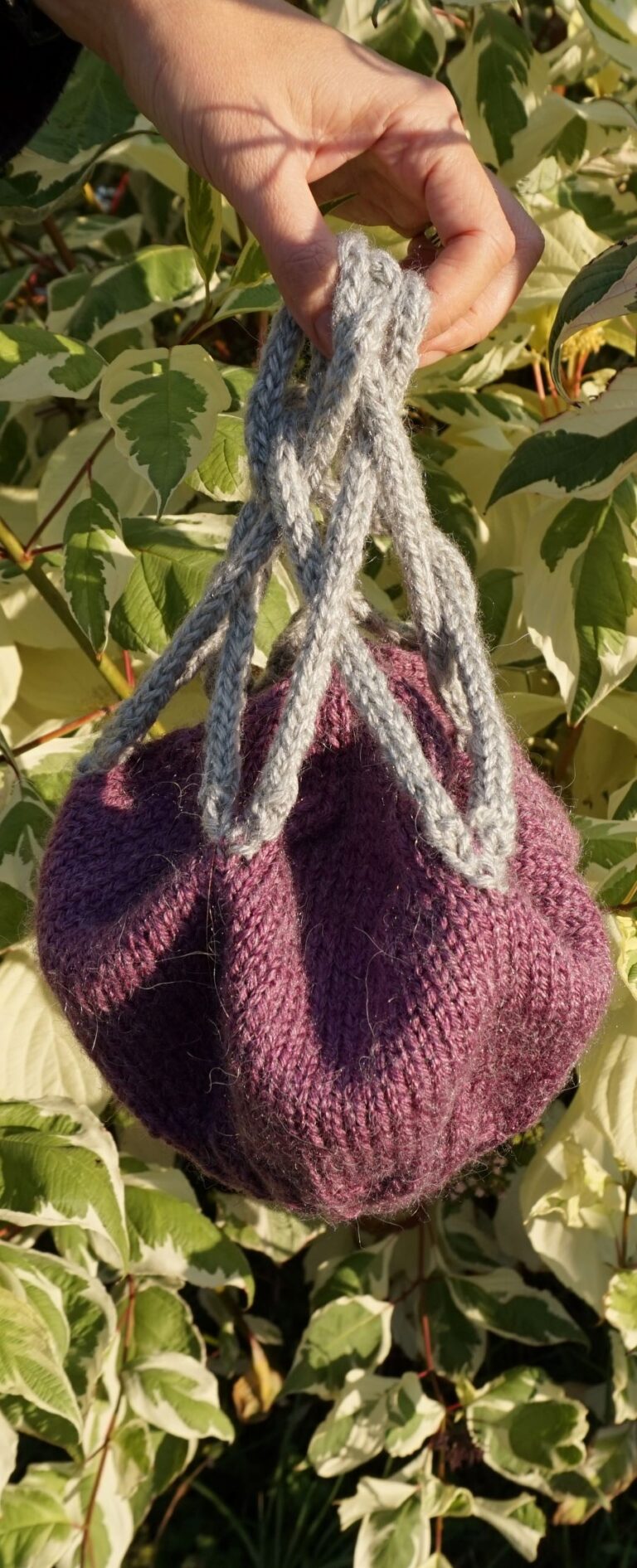 A hand is dangling a grey and purple hat by i-cord strands in front of leaves. -cood strands that form a celtic knot. In