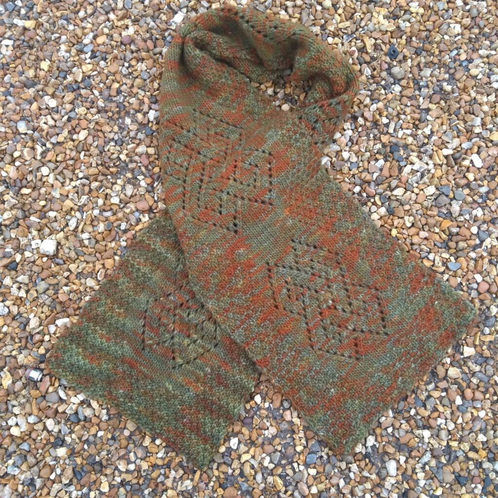 a lace scarf in greens, oranges and browns twisted about itself on a pebble beach.