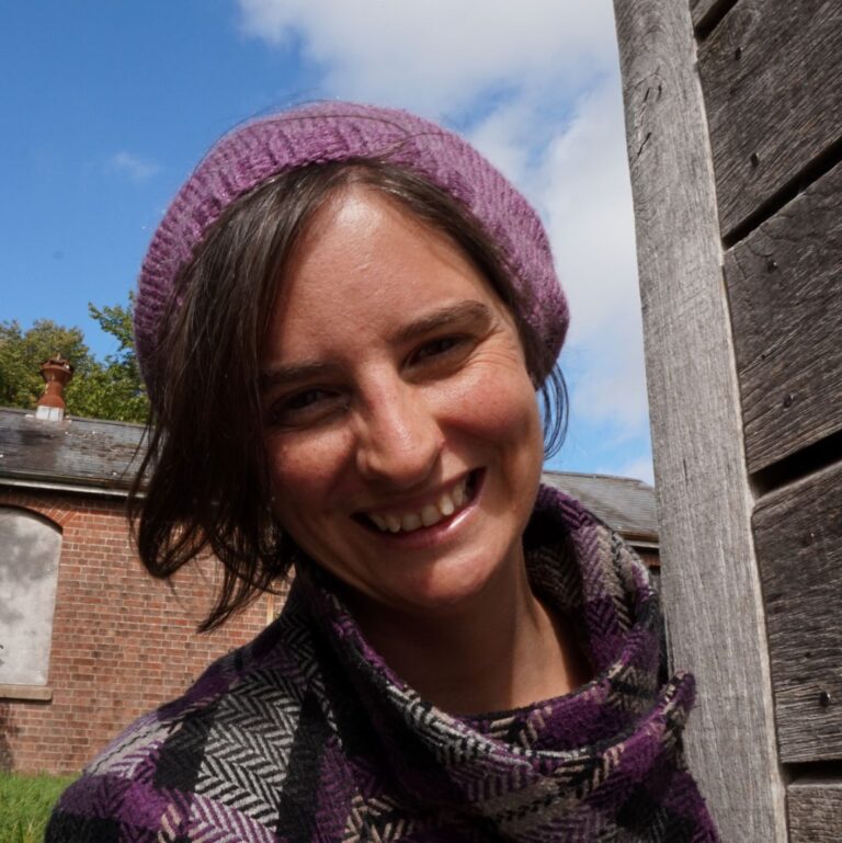 A woman is facing the camera, leaning out from a wall wearing a purple beret with a grey six pointed celtic knot on it formed from i-cords.