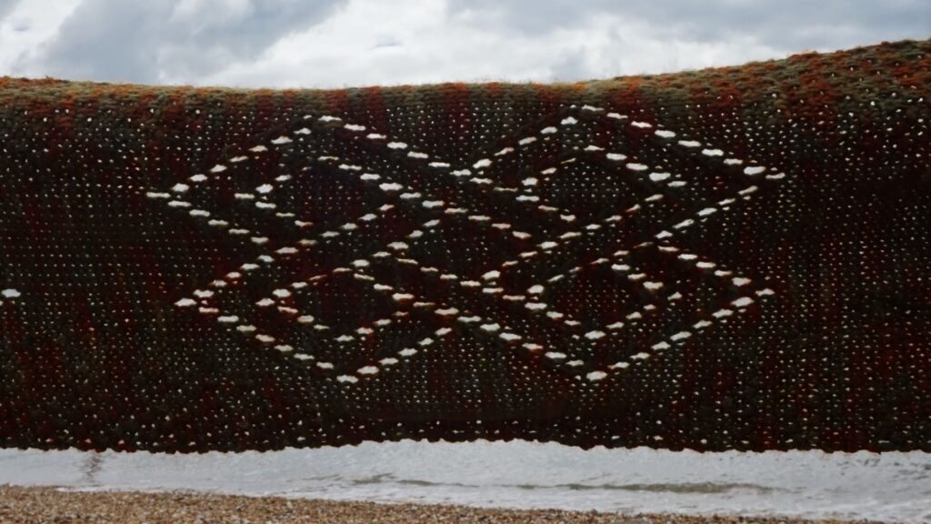 a lace scarf in held against the light to show the lacework celtic knot pattern, a beach and sea can be seen in the background.