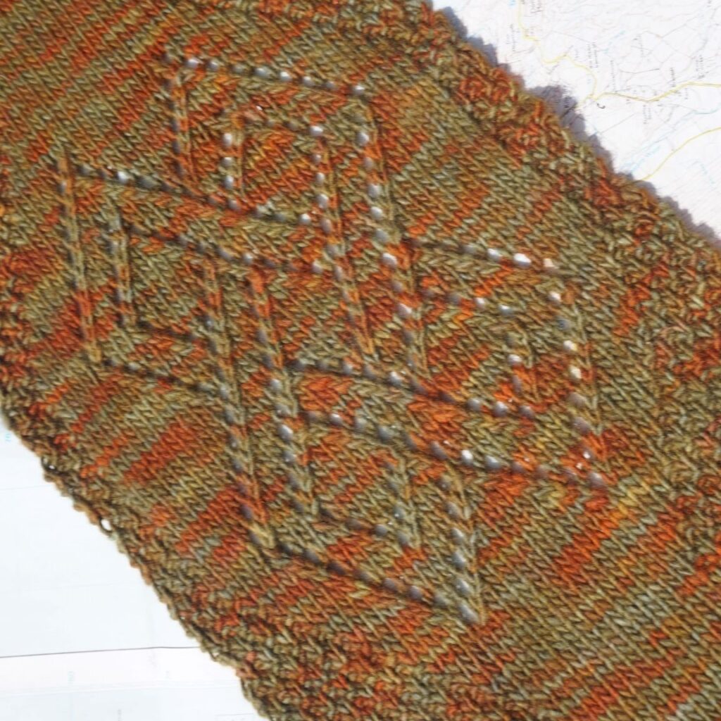 a close up picture of a knitted scarf with a celtic knot worked in openwork lace pattern with a moss stitch border. the scarf is in greens, oranges and browns.