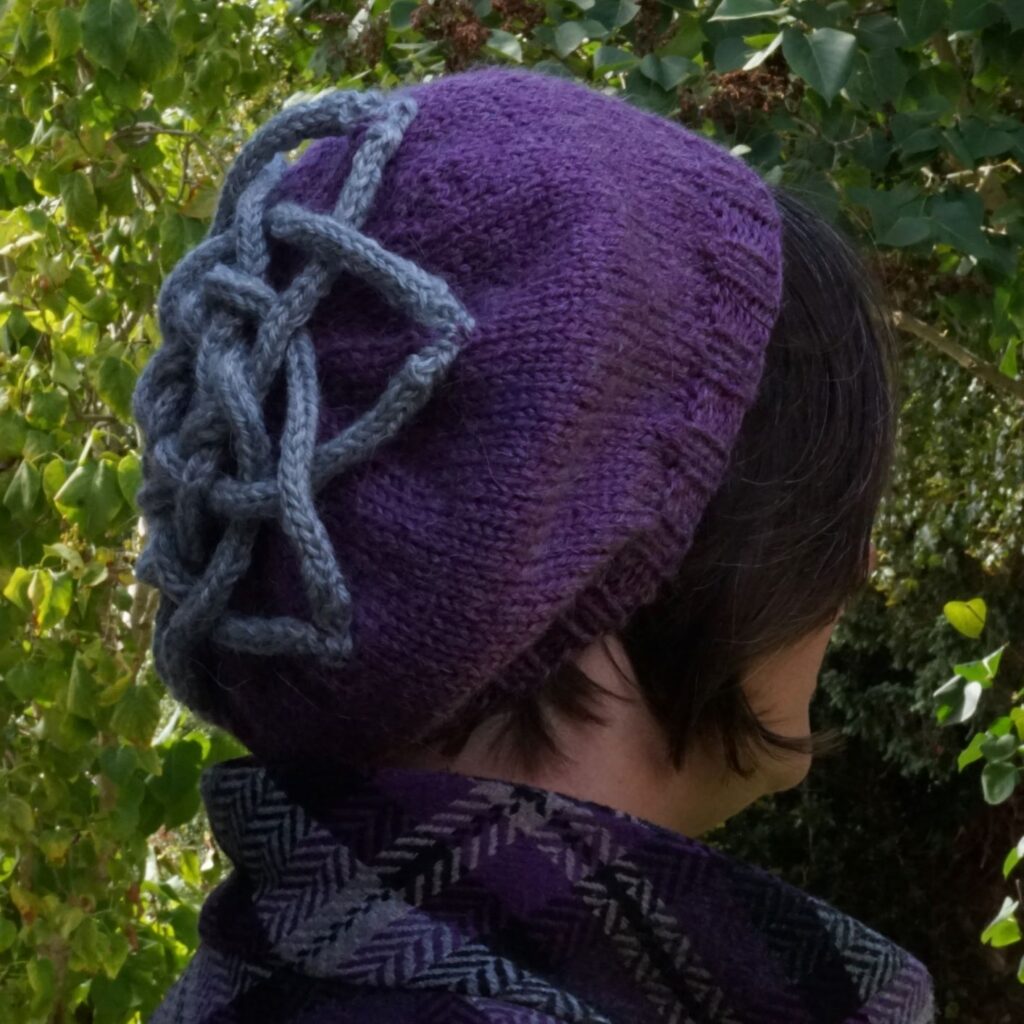 A woman is facing away from the camera, showing the side and back of a purple beret with a grey six pointed celtic knot on it formed from i-cords.