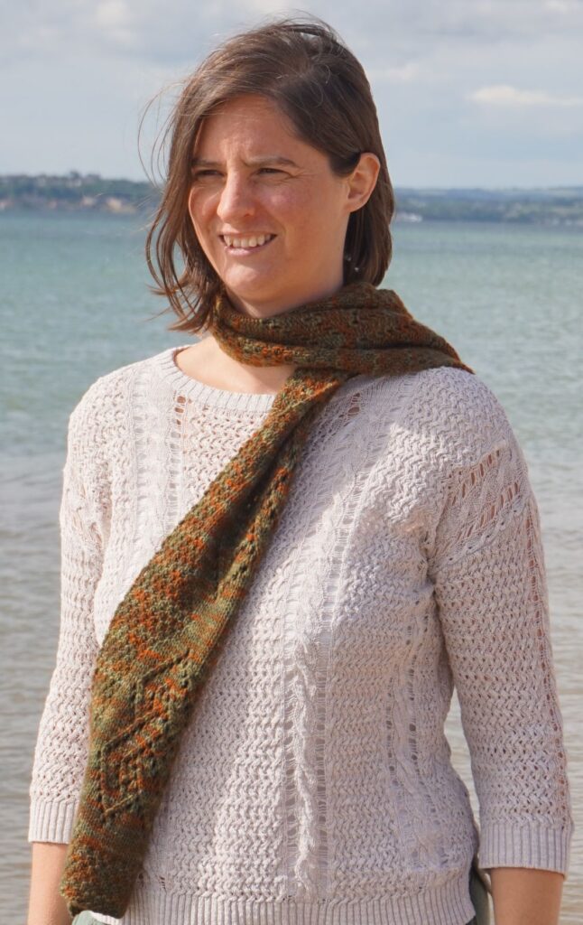 a woman is standing on the edge of the sea wearing a lace scarf in autumn colours