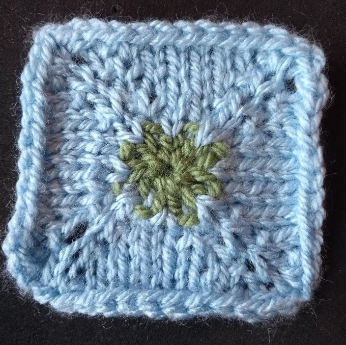 Close up view of a knitted swatch in the shape of a square. The swatch is worked in blue, the pin-hole cast on is worked in green at the centre.