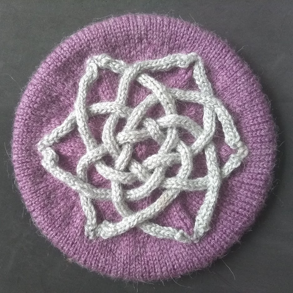a purple tam hat on a black background. the crown is decorated with a celtic knot made from grey i-cord.