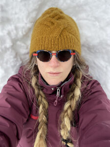 A woman lies on snow wearing a yellow cabled hat.
