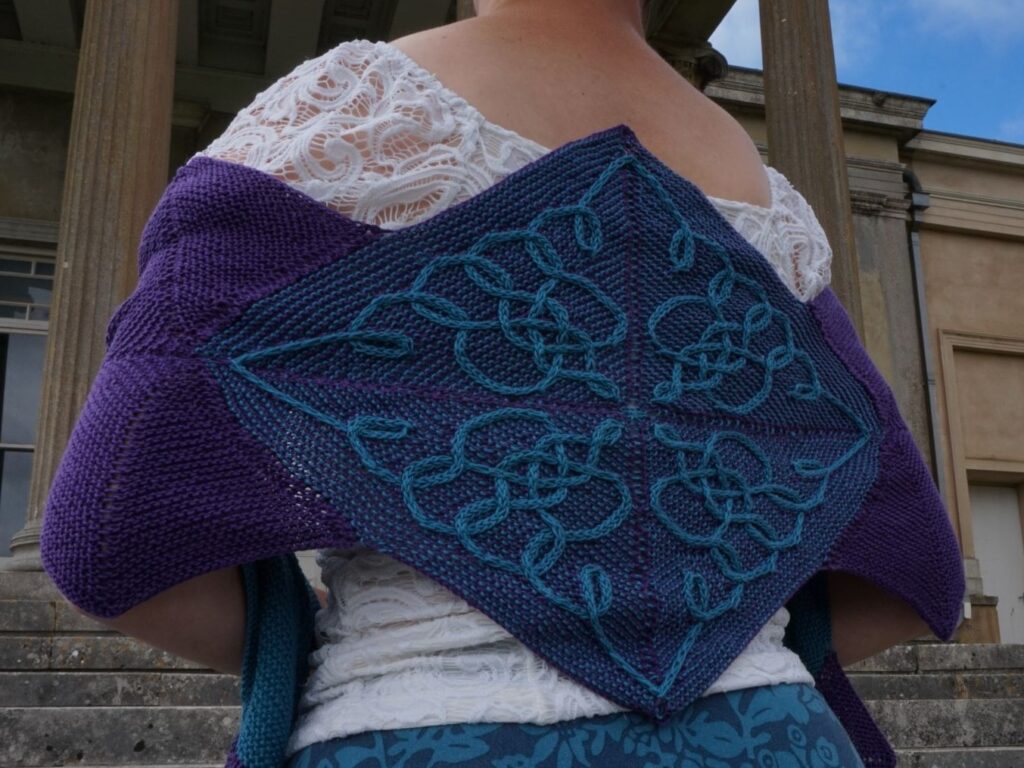 Back view of a diamond shaped wrap spread across a woman's shoulder. The wrap has an intricate cable pattern in purple and green.