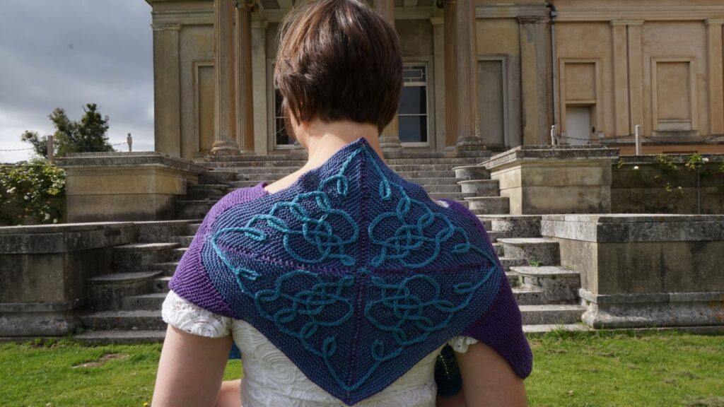 Back view of a diamond shaped wrap spread across a woman's shoulder. The wrap has an intricate cable pattern in purple and green.