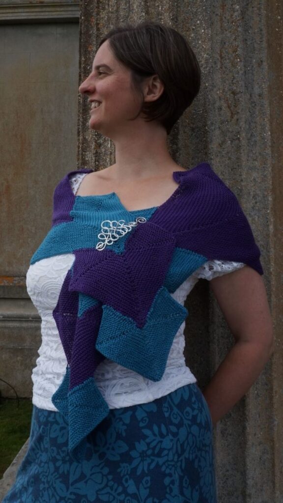 Side view of woman leaning against a pillar wearing a purple and green wrap held closed with a silver brooch.