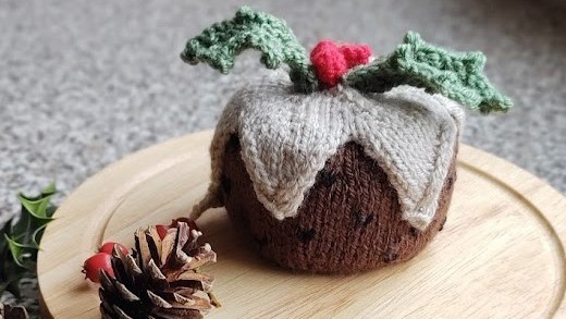 a knitted christmas pudding sits on a wooden board surrounded by pinecones and holly leaves.