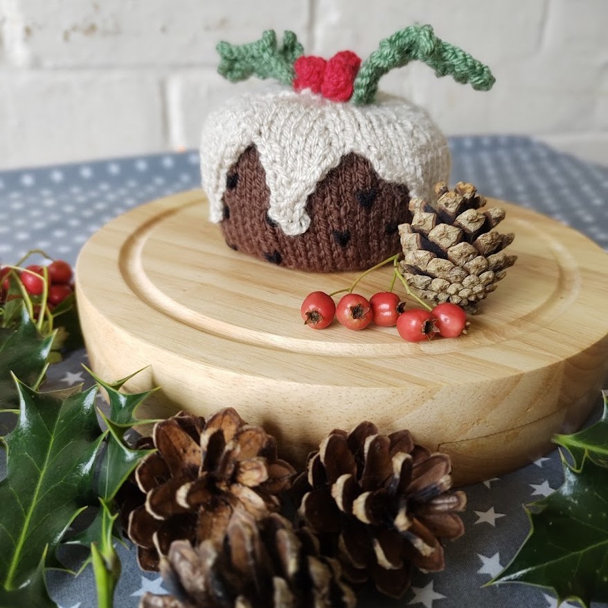 a knitted christmas pudding sits on a wooden board surrounded by pinecones and holly leaves.