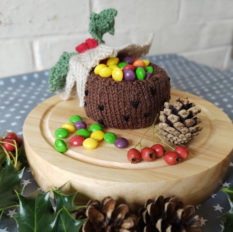 a knitted christmas pudding sits on a wooden board surrounded by pinecones and holly leaves, the top of the pudding is tipped back to reveal sweets and candy inside.