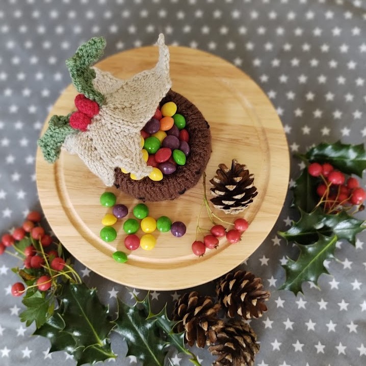 a knitted christmas pudding, viewed from above sits on a wooden board surrounded by pinecones and holly leaves, the top of the pudding is tipped back to reveal sweets and candy inside.