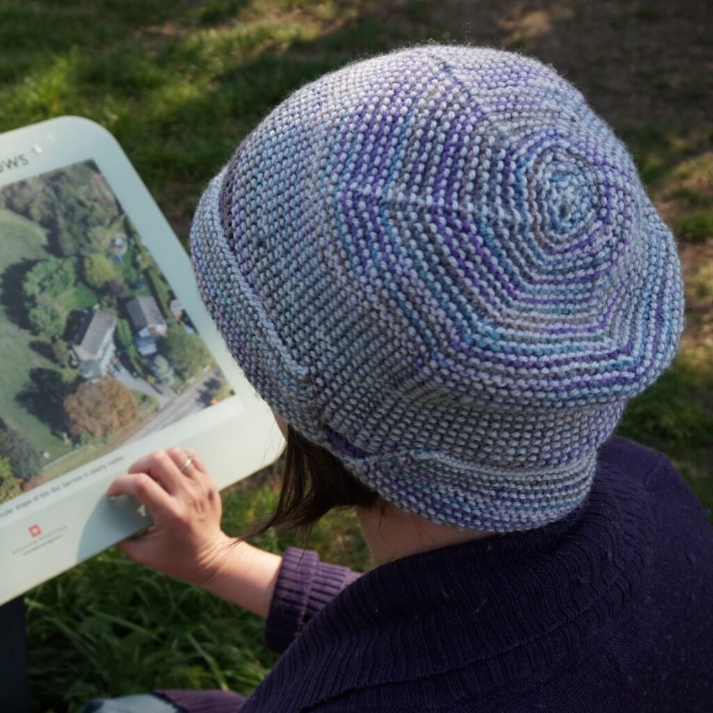 the camera looks down on a woman wearing a silver and purple cloche hat with the crown and brim visible to the camera. She is reading an information sign.