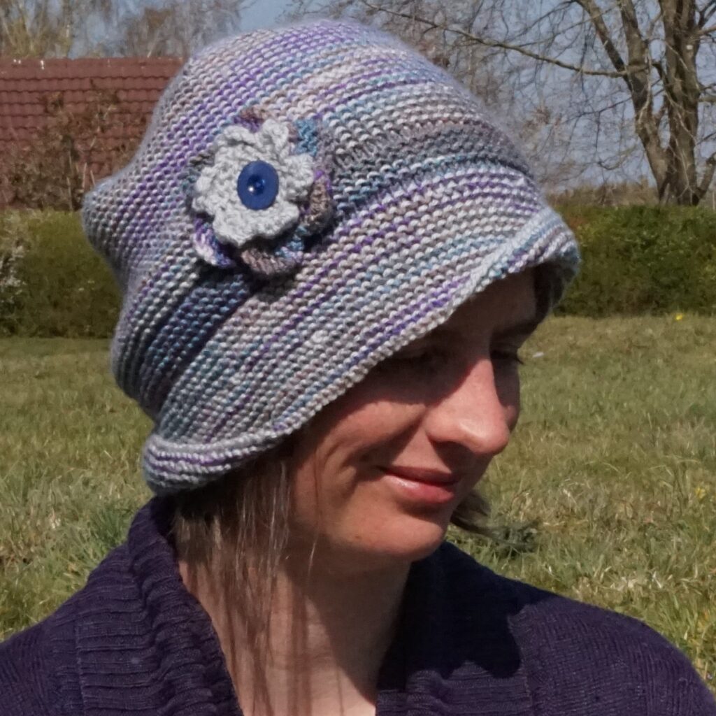 A woman sits in the sunshine in a field with a hedge and trees in the background. She is wearing a purple and grey cloche style hat with assymetrical brim and flower decoration.