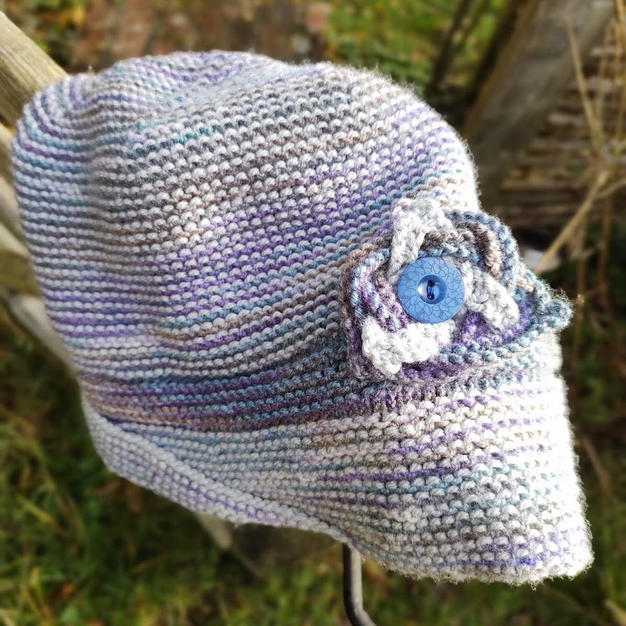 A purple and silver hand knit hat sits on a gate post. it has a celtic knot attached to the hat with a button.