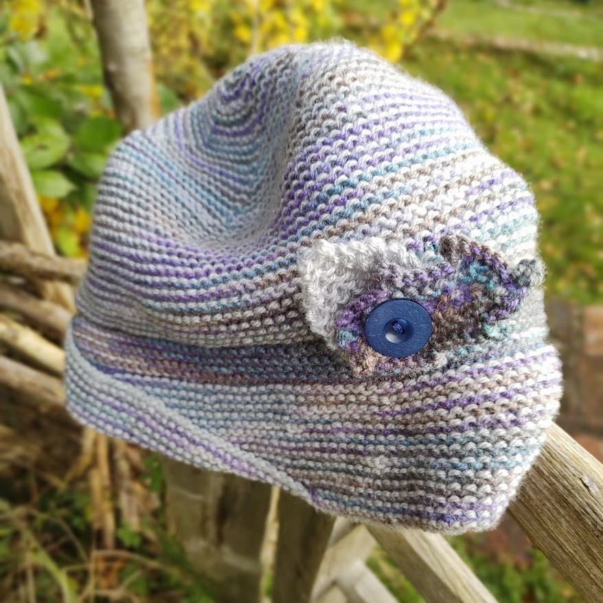 A purple and silver hand knit hat sits on a gate post. it has decorative leaves attached to the hat with a button.