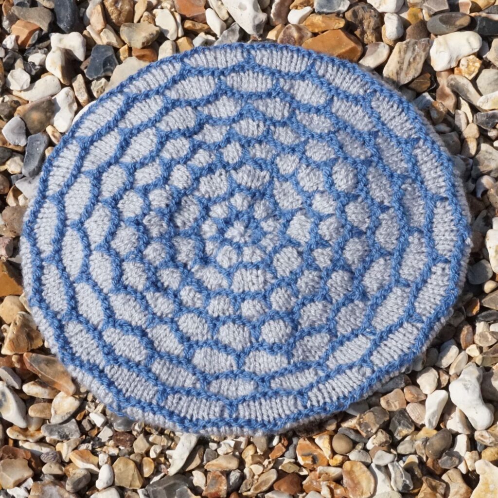 a hat crown is laid on a shingle beach. The crown has decreasing circles of light grey enclosed by blue lines