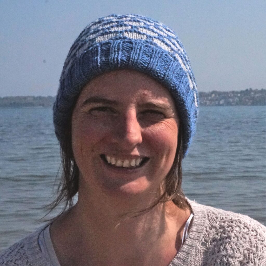 a woman faces the camera with the sea behind her. She wears a hand knitted blue and grey beanie with a deep brim
