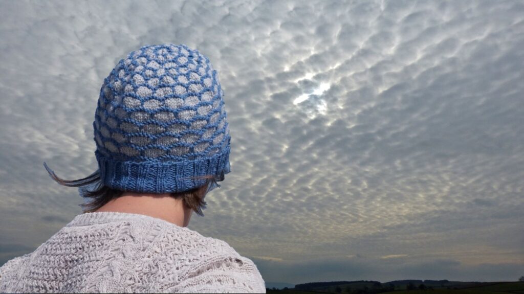 A woman faces away from the camera wearing a blue and grey handknitted beanie hat. The sky in the background has stunning cloud formations.