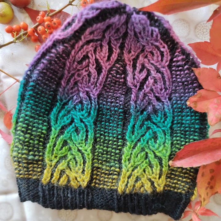 A slightly pointed hat lies flat on a neutral background. The hat is black with rainbow cables worked in brioche stitch,separated by garter-stitch.