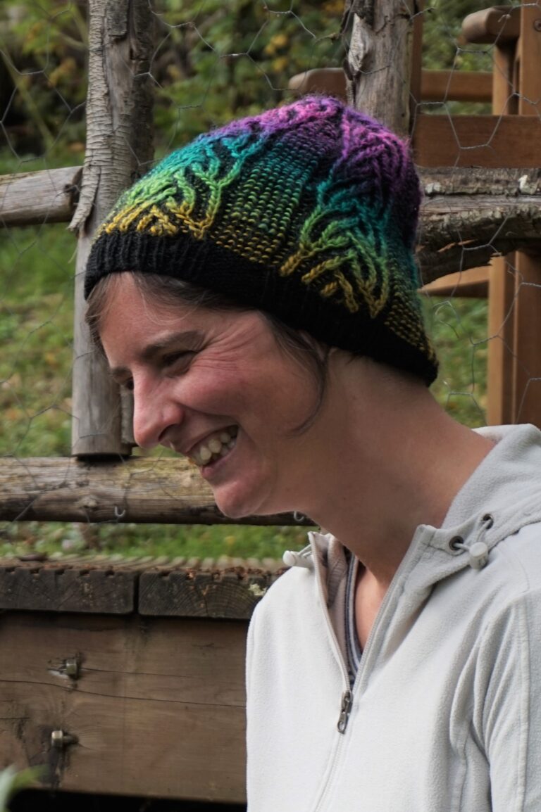 A woman sits next to some garden decking. She is wearing a hand knitted hat with brightly coloured brioche cables running from brim to crown.