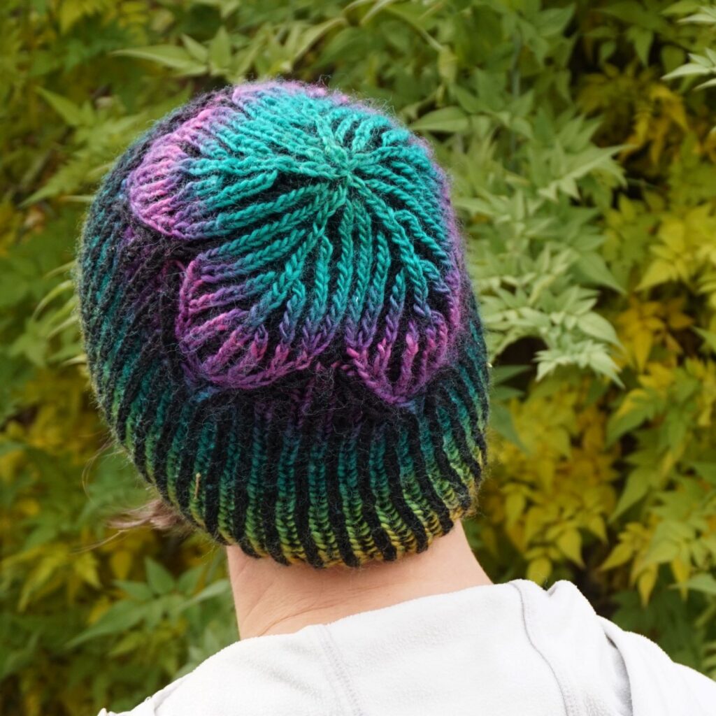 a woman is facing away from the camera, looking towards a leafy hedge. She is wearing a rainbow and black striped hat, with a large star on the crown.