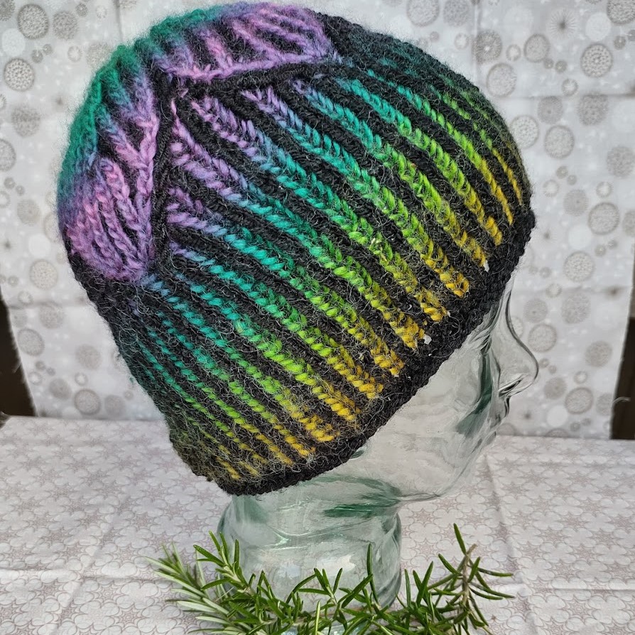 a brightly coloured rainbow striped hat is placed on a glass head. the hat has brioche stripes alternating between bright colours and black, and has a star design on the crown.