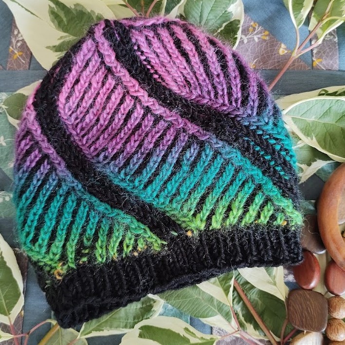 A rainbow coloured hat is placed flat on variegated leaves. It has black stripes running diagonally from the bottom to the top.