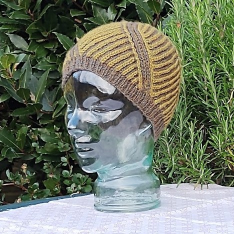 A glass head sits on a table in front of a dark hedge. It is wearing a yellow striped brioche hat with brown brim, and a brown stripes moves diagonally across the hat.