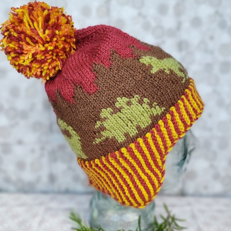 side view of a child's bobble hat. the stegosaurus and t-rex motif can be seen