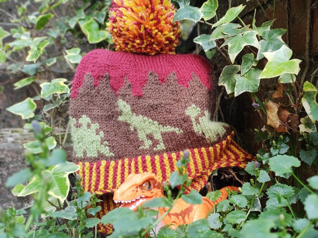 a child's bobble hat with dinosaurs lays on some ivy, an orange velociraptor toy is in the foreground