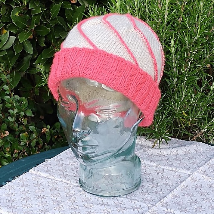 A hat is modelled on a glass head. it is cream with a large pink brim, and swirling pink lines from the brim to crown.